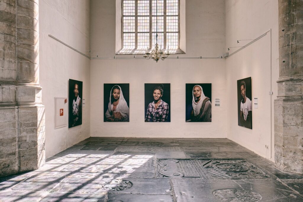 Een galerieruimte met stenen vloeren en hoge ramen toont vijf grote portretfoto's van verschillende personen op een witte muur, verlicht door natuurlijk licht dat door het raam naar binnen valt.