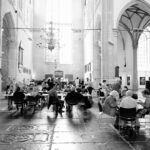 Mensen zitten aan tafels in een grote, lichte zaal met hoge gewelfde plafonds, boogramen en stenen zuilen. De ruimte lijkt op een kerk of een historisch gebouw. Sommigen eten of praten in een ongedwongen, gemeenschappelijke sfeer.