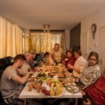 Een groep mensen, waaronder mannen, vrouwen en kinderen, zit rond een tafel met verschillende gerechten, fruit en drankjes in een warm verlichte eetkamer en deelt samen een maaltijd.