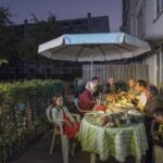 Een gezin verzamelt zich 's avonds buiten rond een tafel en deelt een maaltijd onder een grote parasol. Er staan borden met eten op tafel en een meisje zit iets apart van de groep en kijkt naar de camera.
