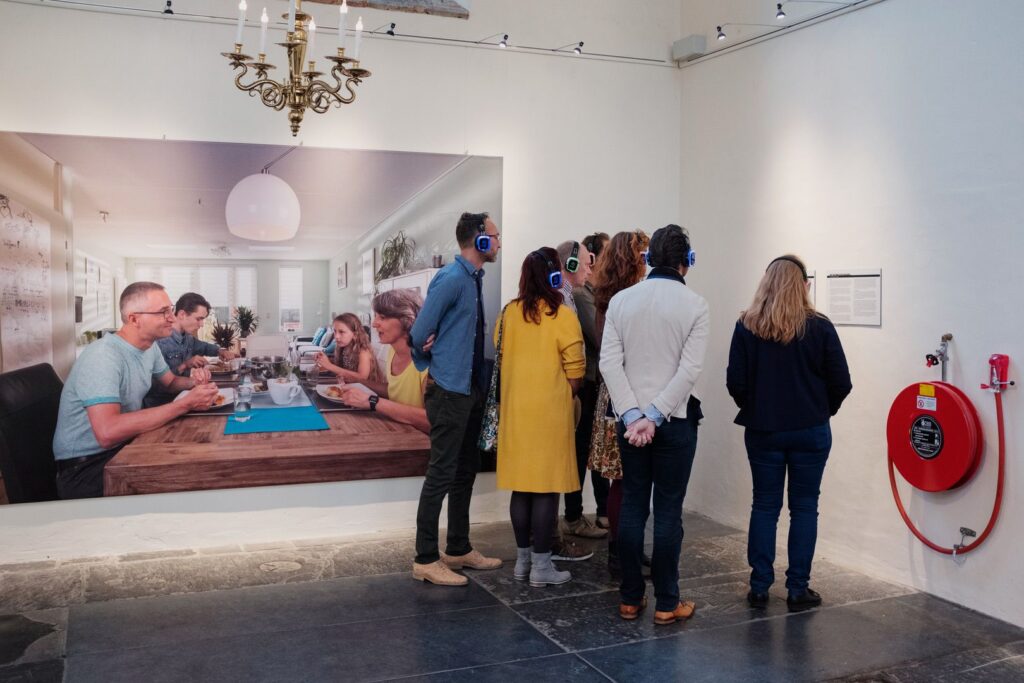 Een groep mensen met koptelefoons staat dicht op elkaar in een kunstgalerie, met hun gezicht naar een muurtekst. Achter hen hangt een grote foto van een gezin rond een eettafel. Op de rechtermuur is een rode brandslang zichtbaar.