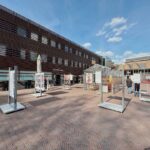 Een buitenplein met kunstdisplays op stands en een kasconstructie, voor een modern gebouw met horizontale lamellen. Een paar mensen lopen rond onder een blauwe lucht met verspreide wolken.