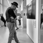 Een man met een hoed en vest staat nadenkend voor een portret in een kunstgalerie. Andere bezoekers staan verspreid op de achtergrond en bekijken verschillende kunstwerken. De omgeving is helder en modern. De afbeelding is in zwart-wit.