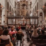Een groot publiek zit in een historische, gewelfde kerk met stenen zuilen en kijkt uit op een podium waar verschillende mensen onder kroonluchters aan een lange tafel zitten. Eén bezoeker zit in een rolstoel.