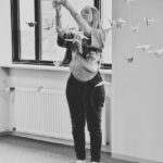 Een vrouw staat binnen en reikt omhoog om de papieren kraanvogels die aan het plafond hangen bij te stellen. De scène is zwart-wit en er valt natuurlijk licht door de grote ramen achter haar.