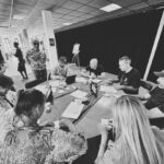 Een groep mensen zit en staat rond een ronde tafel, bezig met een discussie of een activiteit met papieren, notitieboekjes en kopjes. De scène vindt binnenshuis plaats in een goed verlichte kamer. De foto is in zwart-wit.