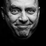 Zwart-wit close-up portret van een oudere man met kort haar, die rechtstreeks naar de camera kijkt en licht glimlacht. De achtergrond is donker, wat zijn gelaatstrekken en uitdrukking benadrukt.