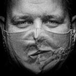 Een close-up, zwart-wit foto van een man die een gezichtsmasker draagt met daarop de onderste helft van een gezicht, waardoor een illusie wordt gecreëerd. De ogen van de man kijken recht in de camera tegen een donkere achtergrond.