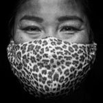 Een close-up zwart-witfoto van een persoon die een gezichtsmasker met luipaardprint draagt, waarbij alleen de ogen en het bovenlichaam zichtbaar zijn, en die rechtstreeks naar de camera kijkt tegen een donkere achtergrond.
