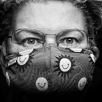 Een persoon met een bril draagt een gezichtsmasker met patroon versierd met lachende zonnen. Het close-up zwart-wit portret benadrukt expressieve ogen en krullend haar.