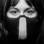 Een close-up, zwart-wit portret van een persoon met lang haar die een gezichtsmasker draagt dat zijn neus en mond bedekt, en met een serieuze uitdrukking recht in de camera kijkt.