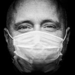 Close-up zwart-wit portret van een persoon met een gezichtsmasker op, waarbij alleen de ogen en het bovenlichaam zichtbaar zijn. Het beeld benadrukt details van de huid en het masker tegen een donkere achtergrond.