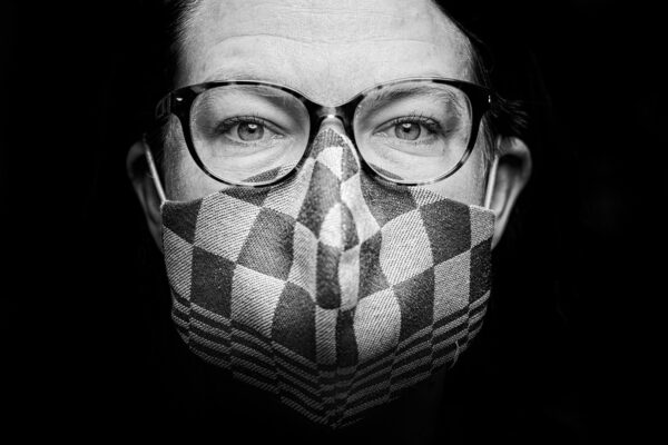 Zwart-wit close-up van een persoon met een bril en een gezichtsmasker met patroon, die rechtstreeks naar de camera kijkt, met een donkere achtergrond.