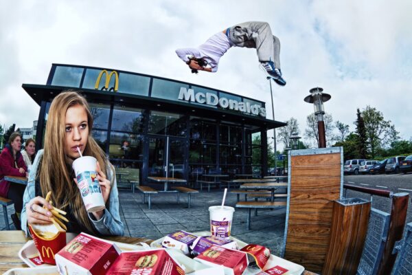 Een jonge vrouw zit aan een tafeltje buiten McDonald's, eet frietjes en nipt van een drankje, terwijl een persoon op de achtergrond een dramatische backflip uitvoert in de lucht boven het zitgedeelte buiten.
