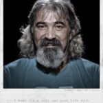 Een man van middelbare leeftijd met lang grijs haar en een baard glimlacht zachtjes naar de camera. Onder het portret staat de tekst: "Ik wens een veilig en goed leven met mijn familie in Nederland.".