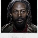 Een man met dreadlocks en een baard kijkt recht in de camera. Hij draagt een camouflagejack en een kastanjebruin overhemd. De achtergrond is zwart. De tekst eronder luidt: "Ik wil vrij zijn", met details: Diallo, 30 jaar, Guinee.