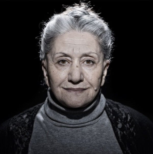Een oudere vrouw met grijs naar achteren gebonden haar, draagt een grijze coltrui en donkere trui, kijkt met een kalme uitdrukking recht in de camera tegen een zwarte achtergrond.