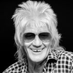 Glimlachende oudere persoon met licht piekhaar en zonnebril, die een zwart-wit geruit overhemd draagt, geposeerd tegen een zwarte achtergrond.