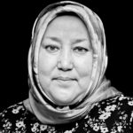 Zwart-wit portret van een vrouw van middelbare leeftijd met een gedessineerde hijab en een gebloemd topje, recht in de camera kijkend met een zachte, neutrale uitdrukking tegen een effen zwarte achtergrond.