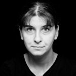 Zwart-wit portret van een persoon met steil donker haar en pony, draagt een effen donker shirt, kijkt recht in de camera tegen een effen zwarte achtergrond.