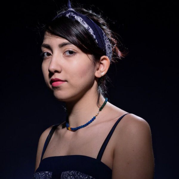 Een jonge vrouw met donker haar gebonden in een bandana kijkt zelfverzekerd naar de camera. Ze draagt een mouwloos topje en een kralenketting, tegen een donkere achtergrond met dramatische belichting.