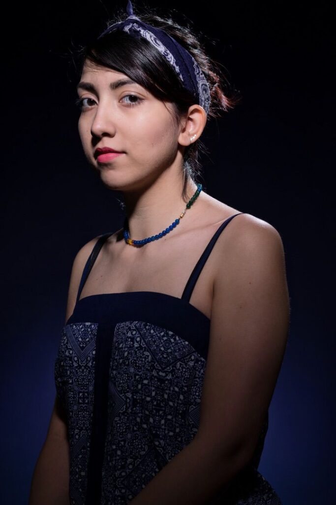 Een jonge vrouw met donker haar en een gedessineerde hoofdband staat tegen een donkere achtergrond. Ze draagt een jurk met blauw patroon en een kralenketting en kijkt met een neutrale uitdrukking een beetje opzij.