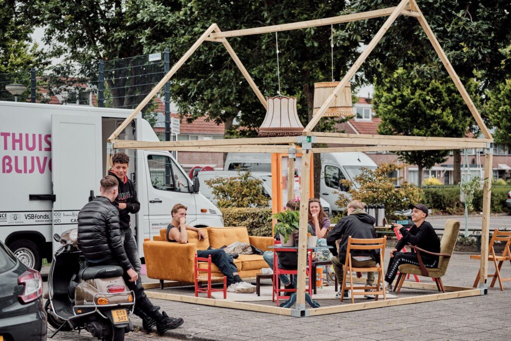 Verschillende mensen zitten en kletsen op kleurrijke banken en stoelen onder een houten frame in de vorm van een huis, dat buiten staat opgesteld in de buurt van geparkeerde voertuigen en groen. Er hangt een ontspannen, gemeenschappelijke sfeer.