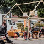 Verschillende mensen zitten en kletsen op kleurrijke banken en stoelen onder een houten frame in de vorm van een huis, dat buiten staat opgesteld in de buurt van geparkeerde voertuigen en groen. Er hangt een ontspannen, gemeenschappelijke sfeer.