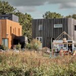 Achter een grasveld staan twee moderne, doosvormige huizen met een houten buitenkant. Aan de voorkant serveren een kleine witte foodtruck en een open houten structuur mensen die op een zonnige dag buiten aan tafels zitten.