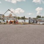 Twee mensen zitten in een open, gedeeltelijk omlijste buitenstructuur bij een rivier, omringd door meubilair en planten, met in de buurt geparkeerde fietsen en industriële gebouwen op de achtergrond onder een gedeeltelijk bewolkte hemel.