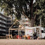 Een witte bestelbus en een open houten constructie staan onder hoge bomen bij een flatgebouw. Verschillende mensen, waaronder kinderen, zitten of spelen rond de opstelling, die is versierd met een "BUI"-banner.