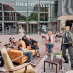 Vijf mensen zitten en staan vrolijk rond ouderwets meubilair op een buitenplein, lachen en vieren feest voor een gebouw met de tekst "TAQA THEATER VEST". Een fotograaf legt het moment vast.