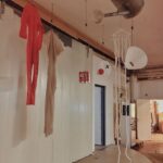 Kunstinstallatie met een rode bodysuit, een transparante beige stof, een metalen gieter en abstracte witte vormen met touwtjes, allemaal hangend aan het plafond in een industriële binnenruimte.