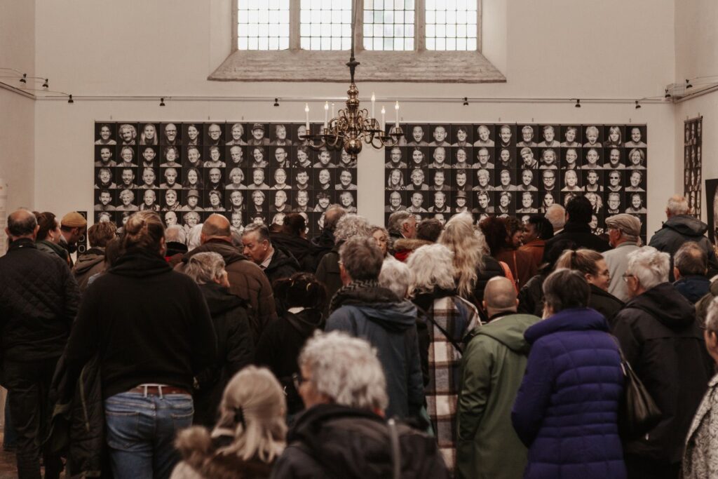 Een grote groep mensen staat binnen en kijkt naar een muur bedekt met een raster van portretfoto's in zwart-wit. Aan het plafond hangt een kroonluchter en grote ramen laten licht binnen boven het scherm.