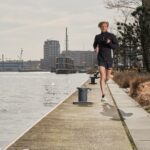 Een persoon in sportkleding jogt over een pad langs het water met gebouwen en droog gras, met op de achtergrond een boot en een stadsgezicht onder een bewolkte hemel.