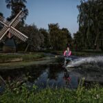 Een persoon waterskiet op een smal kanaal in de schemering, waardoor een nevel van water ontstaat, met een kleine windmolen en weelderige groene bomen op de achtergrond.