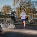 Een man met een grijs t-shirt, een blauwe korte broek en een zonnebril rent op een verhard pad langs een rivier, omringd door grote rotsen en bladerloze bomen op een zonnige dag.