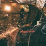 Een arbeider met beschermende kleding gebruikt een haakse slijper op een grote metalen pijp, waardoor felle vonken ontstaan. De omgeving ziet er industrieel uit, met gereedschap, draden en apparatuur zichtbaar op de achtergrond.