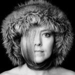 Zwart-wit portret van een persoon met een harige kap. Een deel van het sluike haar valt over één oog en wikkelt zich om de nek. De persoon kijkt recht in de camera tegen een donkere achtergrond.