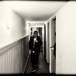 Een persoon met pet en jas loopt door een smalle gang met witte muren en deuren aan de rechterkant; een andere persoon is gedeeltelijk zichtbaar achter een deur. De afbeelding is in zwart-wit.