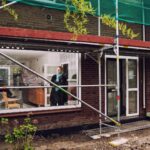 Een persoon staat in een huis en kijkt uit een groot raam. De buitenkant van het huis is omringd door steigers, wat duidt op bouw- of renovatiewerkzaamheden. De tuin heeft kale grond en verspreide bakstenen.