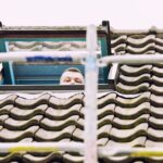 Het gezicht van een persoon is gedeeltelijk zichtbaar, glurend uit een open dakraam op een pannendak. Steigerbalken doorkruisen de voorgrond en de dakpannen lijken verweerd en bemost.