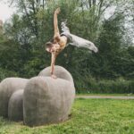 Een shirtloze man voert een eenhandige handstand uit op grote, ronde stenen sculpturen in een graspark met bomen en flatgebouwen op de achtergrond.