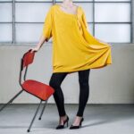 Een persoon met een los geel topje, een zwarte legging en zwarte hakken staat naast een rode stoel en houdt de zijkant van het topje vast. De setting is een minimalistische kamer met grote matglazen ramen.