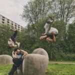 Drie mensen maken acrobatische salto's en sprongen van grote betonnen sculpturen in een graspark, omringd door bomen en een gebouw op de achtergrond. Eén persoon zit terwijl de andere twee midden in de lucht zijn.