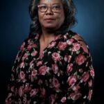 Een vrouw met grijs haar en een bril poseert tegen een donkerblauwe achtergrond, draagt een zwarte gebloemde blouse met roze rozen en houdt haar handen samen in haar schoot.