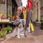 Twee vrouwen lopen arm in arm door een charmant stenen straatje, glimlachend en met boodschappentassen. Ze dragen casual, stijlvolle kleding en een zonnebril. Op de achtergrond zijn bloemstukken en winkelpuien te zien.