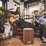Twee mannen zitten op een bruinleren bank en bespelen muziekinstrumenten - de een met een akoestische gitaar, de ander met een basgitaar - naast kamerplanten in een gezellige kamer met grote ramen en een tapijt met patronen.