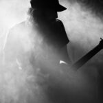 Een silhouet van een gitarist met een pet op, omgeven door rook of mist, speelt op het podium. De scène is sfeervol en dramatisch, met licht dat schaduwen creëert en de contouren van de gitaar benadrukt.