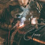 Een arbeider met beschermende kleding en een lasmasker last een grote metalen pijp, waarbij vonken en rook opstijgen uit het laspunt in een industriële omgeving.